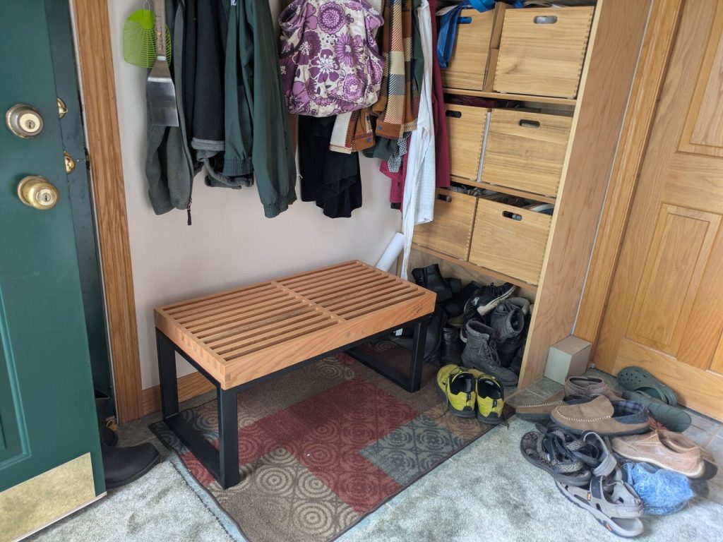 The finished oak and black steel bench in the entry, next to the cubby shelves, with coats hanging above and shoes on the floor